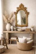 May include: A gold ornate mirror hangs above a rustic wooden console table. The table displays vases, bowls, and decorative items. A woven basket with pillows and a jute rug complete the neutral-toned, bohemian-style decor.