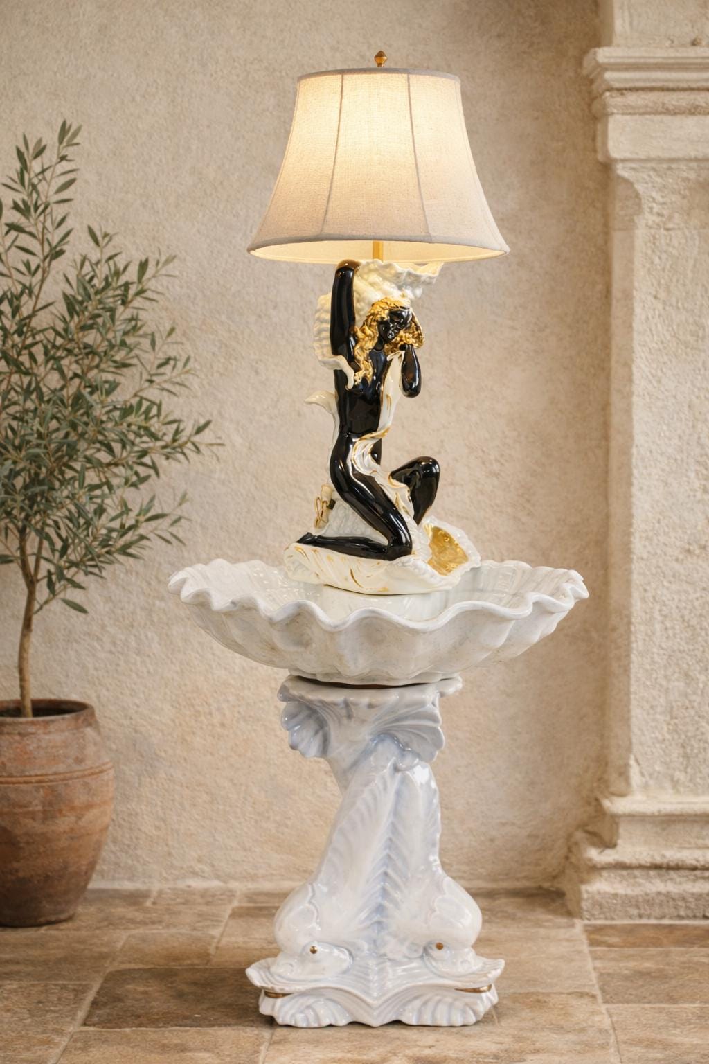 May include: A decorative lamp with a black and gold figure supporting a lampshade. The lamp sits atop a white, shell-shaped basin, mounted on a pedestal with a dolphin design. The lamp is a unique home decor item.