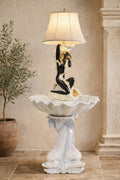 May include: A decorative lamp with a black and gold figure supporting a lampshade. The lamp sits atop a white, shell-shaped basin, mounted on a pedestal with a dolphin design. The lamp is a unique home decor item.